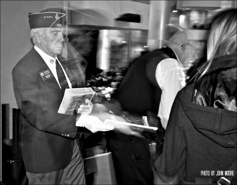 Veteran Richard Callan helps out the ushers by passing out programs to arriving guests moments before the presentation of the Color Guard. Photo by John Moore.  